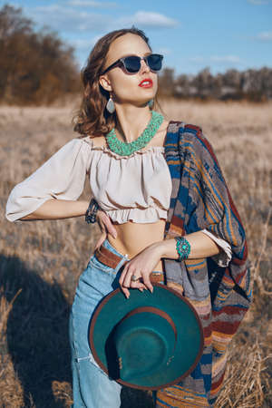 Spirit of freedom. An attractive boho girl in stylish clothes and sunglasses stands in the field in the countryside in warm sunlight. Vacation, traveling. Bohemian, modern hippie style.の写真素材