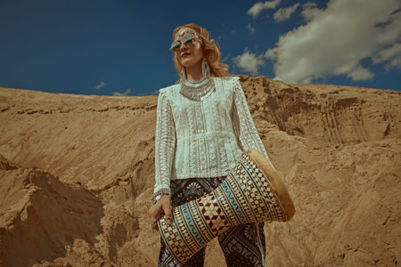 Stylish blonde girl in boho clothes and jewelry stands on the background of sand dunes with a...