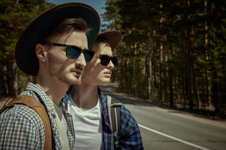 Two handsome men travellers stand together by the road against the backdrop of a pine forest waiting for a passing car. Hipsters outdoors. Hitchhiking, travel and tourism.の写真素材
