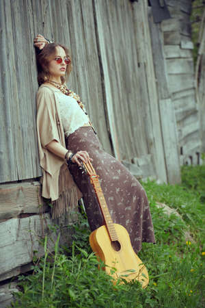Portrait of a beautiful girl model in boho style clothes and jewelry standing with a guitar by an old wooden fence in the countryside. Summer style, vacation. Fashion shot.の写真素材