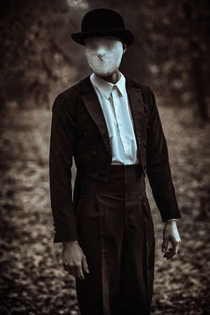A strange and creepy gentleman in a mask of plaster bandage on his face, in a bowler hat and a black tailcoat, stands in an old autumn park. Halloween. Vintage style. Horror, thriller.の写真素材