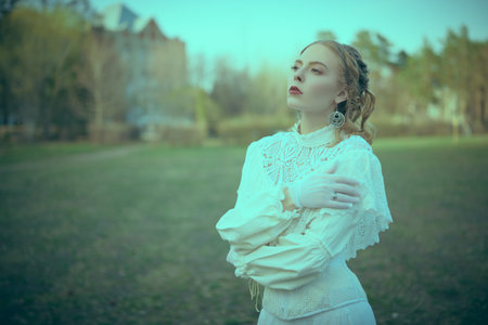 A refined, beautiful aristocratic girl in a white lace dress stands dreamily in the park of the...