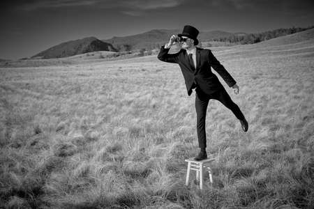 Black-and-white portrait of a man in black suit, top hat and black sunglasses standing on a stool in a valley and looking into the distance through binoculars. Surrealist style.の写真素材