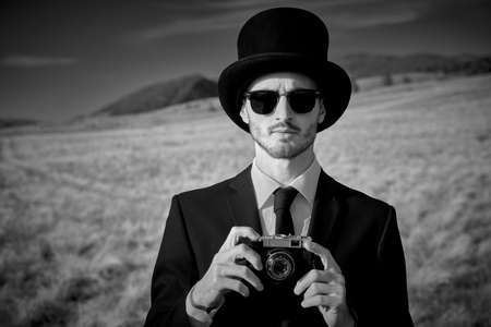 Black and white portrait of an elegant man in black suit, top hat and black sunglasses standing in a valley with an old camera in his hands. Surrealist style.の写真素材