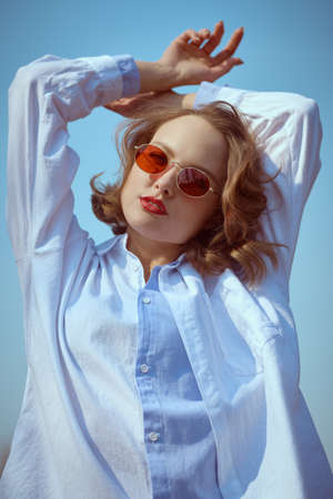 Beautiful brunette woman with elegant makeup, hairstyle and sunglasses wearing a blue shirt stands outdoors on a sunny spring day in the background of blue sky. Makeup, hairstyle and glasses style.の写真素材