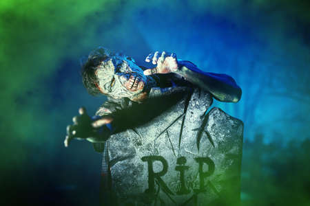 A terrible bloodthirsty zombie man climbs out from behind a tombstone in a cemetery in the light of the moon and surrounded by fog. Halloween, horror.の写真素材