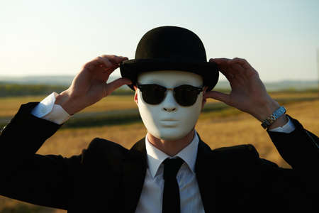 A surreal gentleman in a black suit, a bowler hat and a white mask stands in a valley looking at the camera.の写真素材