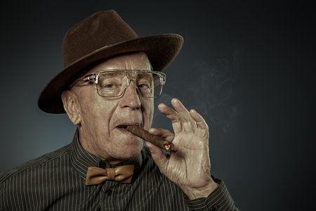 An old gentleman smoking a cigar. Detective story character. Studio portrait on a dark background.の写真素材