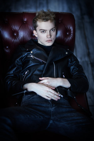 Men's fashion and style. A handsome young man in black leather jacket and black jeans rests on a leather sofa in loft style room.の写真素材