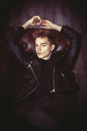 A handsome guy dressed in black leather jacket and black jeans rests on a leather sofa in loft style room. Men's fashion and style.の写真素材