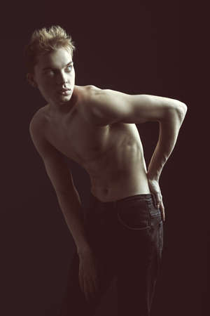 Portrait of a young man in jeans and with a thin naked torso, hunched over from back pain. Black studio background. People and health problems.の写真素材