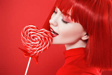Beautiful bright girl with red lips and red shoulder-length hair tries a heart-shaped lollipop. Red studio background. Beauty, cosmetics, makeup. Hairstyle, hair coloring.の写真素材