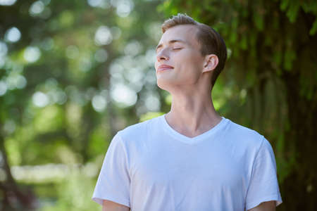 Happy people, positive emotions, lifestyle. A happy guy enjoys a sunny summer day in the park standing with closed eyes.の写真素材