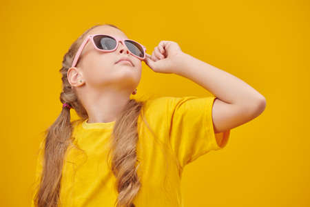 A stylish little blonde girl with pigtails poses in a bright yellow t-shirt and fashionable sunglasses looking up on a yellow background. Children's summer fashion.の写真素材