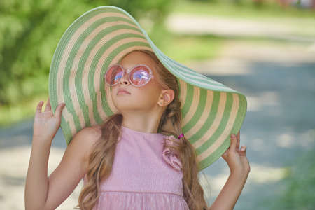 A charming girl child with wavy blonde hair wearing fashionable summer pink dress, stylish sunglasses and wide-brimmed hat walks on a street. Kid's summer fashion. Summer holidays.の写真素材