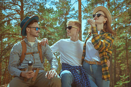 A girl and two guys drink tea from a flask, stopping for a halt on a hike. Summer tourism, hiking. Vacation. Tourist equipment.の写真素材