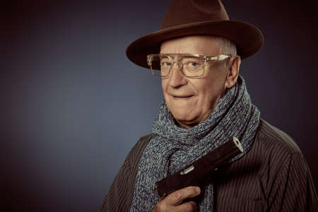 Portrait of a brutal elderly gentleman in hat, glasses and scarf holding a gun in his hand. Detective story character. Studio portrait on a dark blue background.の写真素材