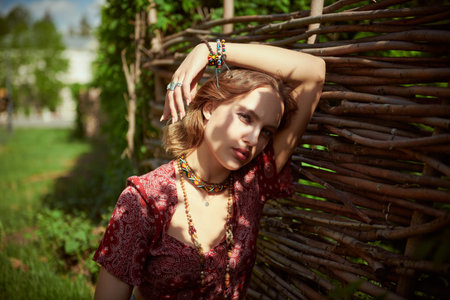 Summer beauty. Beautiful girl in hippie style clothes and jewelry rests by a wicker fence in the countryside. Boho and hippie style, fashion. Vacation.の写真素材