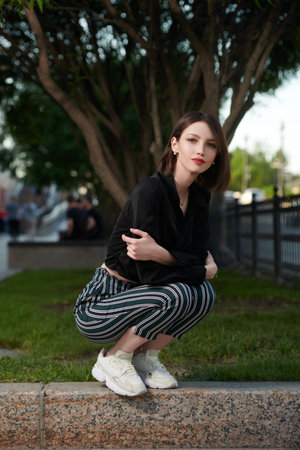 A beautiful young woman with bob haircut in black and white clothes sits on a city street on a nice...