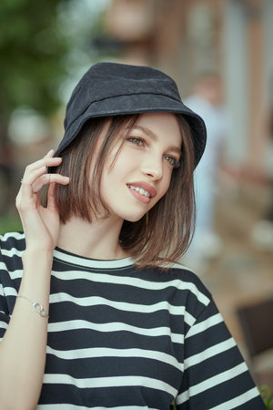 A cute brunette girl with bob haircut in a striped black and white blouse and black bucket hat stands on a city street on a nice summer day and smiles. People concept, lifestyle. Summer vacation.の写真素材