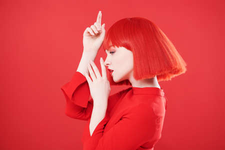 Adorable girl with bright red lips and red hair posing in profile. Red studio background. Beauty, makeup. Hairstyle, hair coloring. Fashionable red colour.の写真素材