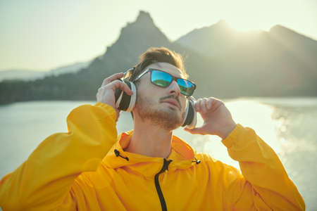 Travel, summer vacations outdoor. A male tourist athlete meets the sunset and listens to pleasant music on headphones, standing on a mountain against a beautiful landscape. Harmony with nature.の写真素材