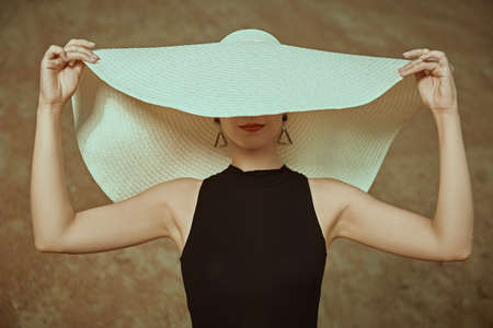 A charming young woman in a black swimsuit and an elegant wide-brimmed hat covering her eyes stands on the beach and smiles. Summer fashion. Vacation at the sea.の写真素材