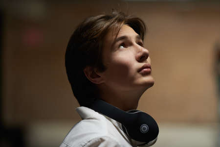 Portrait of a handsome teenage boy with headphones on his neck standing on a city street and looking thoughtfully up. Modern generation, adolescence.の写真素材