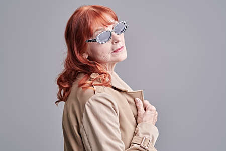 Portrait of an old lady with bright red hair wearing modern clothes and stylish sunglasses. Studio portrait on a grey background. Fashion for the elderly.の写真素材
