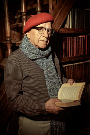 An intelligent old man in a beret and glasses is reading a book while standing at home with a classic vintage interior.の写真素材