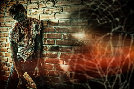 Zombie man. A bloodied zombie stands leaning against a brick wall in a dark alley of a city destroyed by the apocalypse. Halloween. Horror movie.の写真素材