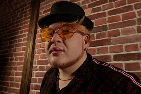 A cool plump guy in a casual sports suit, black hat and yellow round glasses stands in a loft style...