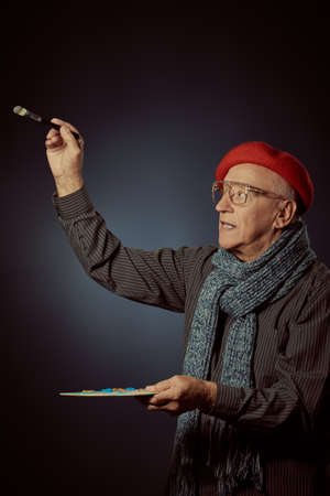 A creative elderly artist in a stylish beret, scarf and glasses creates a masterpiece with the help of his palette and a brush. Studio portrait on a dark blue background.の写真素材