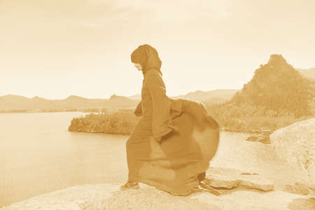 A male wanderer in a black hooded robe resists a strong wind while standing on a mountaintop. Beautiful mountain landscape background. Fantasy and adventure.の写真素材