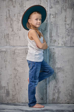 Portrait of a fashionable little girl in a western style hat, white T-shirt and jeans posing against a grunge wall background. Children's fashion. Full length shot.の写真素材