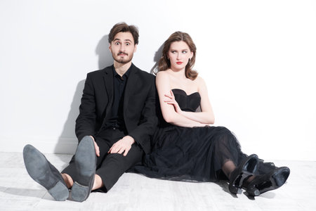 A loving couple in elegant black evening clothes sitting on the floor next to each other with...