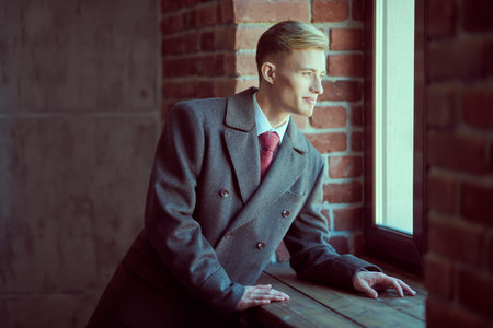Young handsome man in elegant coat, white shirt and tie looks out the window in a loft style apartment. Men's fashion and beauty. Lifestyle.の写真素材