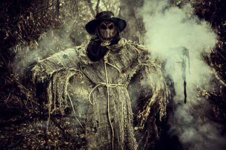 Halloween. An evil terrible Scarecrow with a bag on his head, in a sackcloth vestment and a black hat, stands in the thicket of the forest, surrounded by fog. Horror, thriller.の写真素材