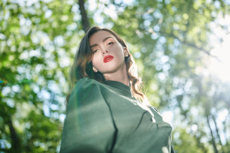 A beautiful girl with wavy dark hair stands under the canopy of trees in a summer park. Sun glare...