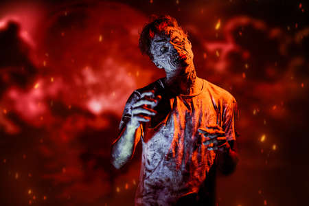 A scary bloodied zombie stands against the background of red clouds of smoke and fire. The Apocalypse. Halloween, horror movie.の写真素材