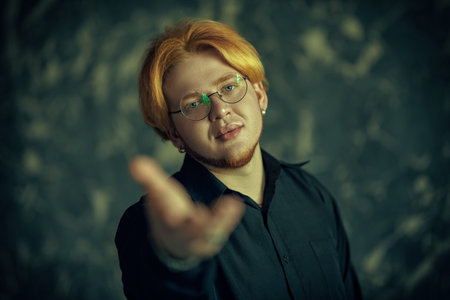 A handsome young plump man with red hair wearing a black shirt and glasses holds out his hand to the...