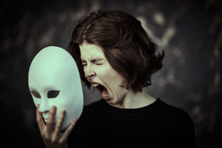 A suffering girl in black clothes screams with all her might holding a mask in front of her. Dark grunge background. Human roles. Hypocrisy. Mental disorders.の写真素材