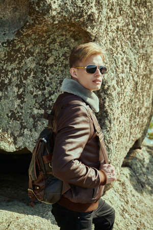 Adventure and travel. A handsome young man in a leather jacket and a backpack behind his back stands by a large rock. Leather fashion.の写真素材