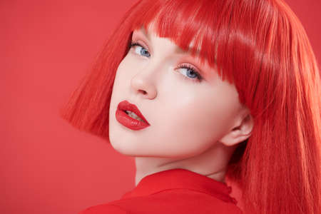 A charming girl with bright red lips and red hair. Red studio background. Beauty, makeup. Hairstyle, hair coloring. Fashionable red colour.の写真素材