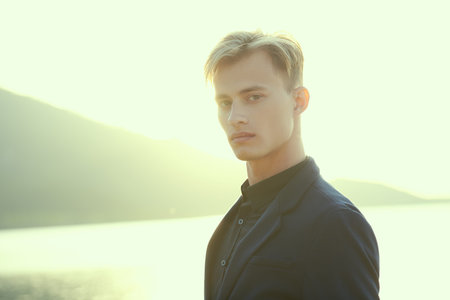 A handsome blond man in black suit stands on the sea coast in warm rays of the setting sun.