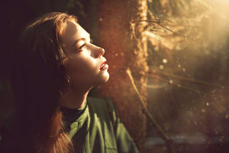 A lovely girl in a green dress stands in a dark enchanted forest looking at the magic light. Legends and fairy tales. Fantasy world. Copy space.の写真素材