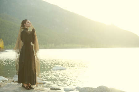 Young beautiful woman in an elegant black 19th century dress stands under the veil by the sea at sunset joining her hands in a prayer and looking with hope into the distance. 19th century novel.の写真素材