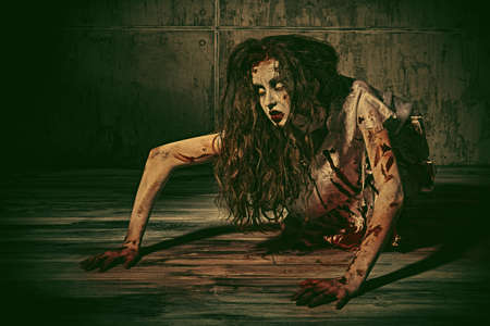 A creepy, devil-possessed woman with pale eyes and tousled hair covering her face and a bloody nightie crawls against a concrete wall. Zombie woman. Horror, thriller. Halloween.の写真素材