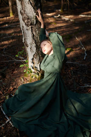 Portrait of a fairy tale girl in a long green dress sitting by a tree in a thick enchanted forest. Fantasy, fairy tale heroine.の写真素材