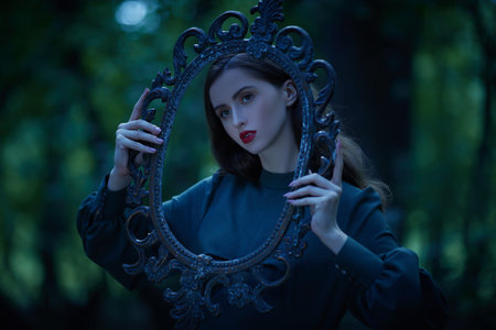 A charming fairy tale girl with dark wavy hair standing in a dark forest and looking through a magical frame. Fantasy, the heroine of a fairy tale.の写真素材